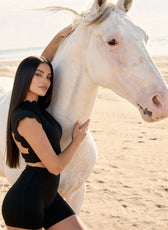 Woman in black outfit standing next to a white horse on a beach