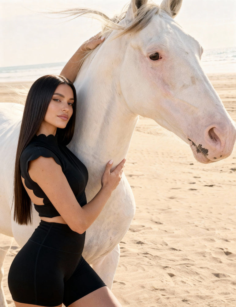 Woman in black outfit standing next to a white horse on a beach