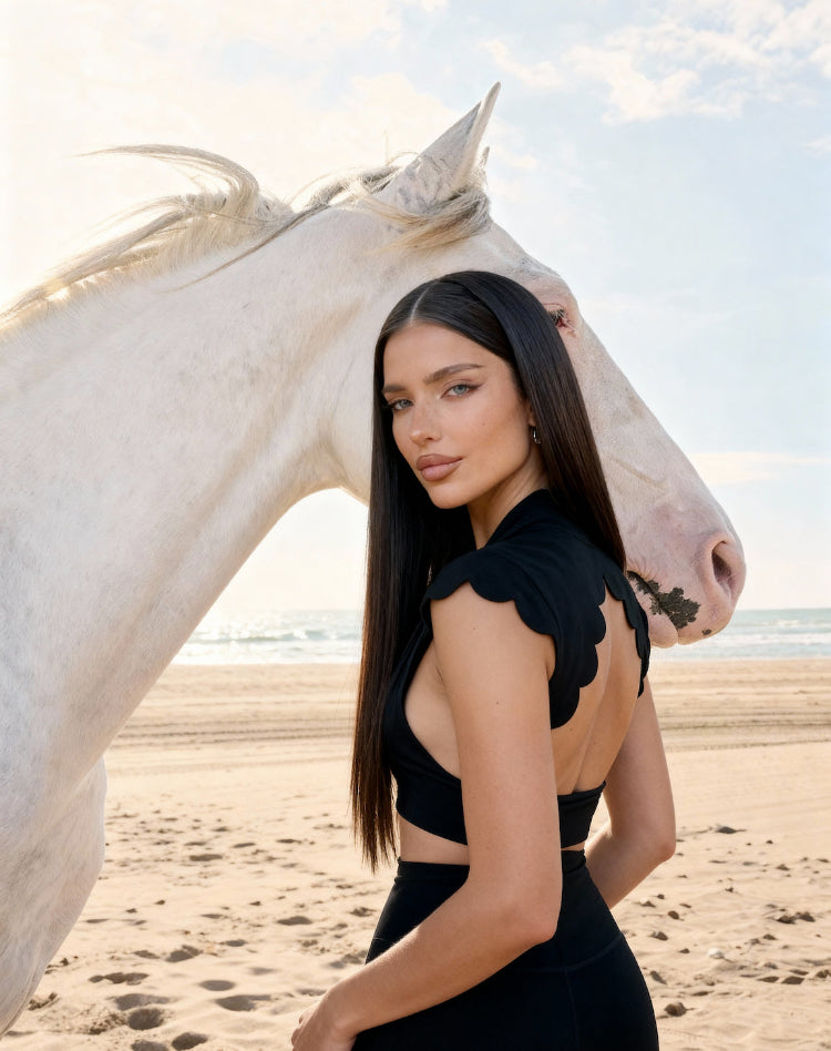 Woman in a black dress standing next to a white horse on a beach