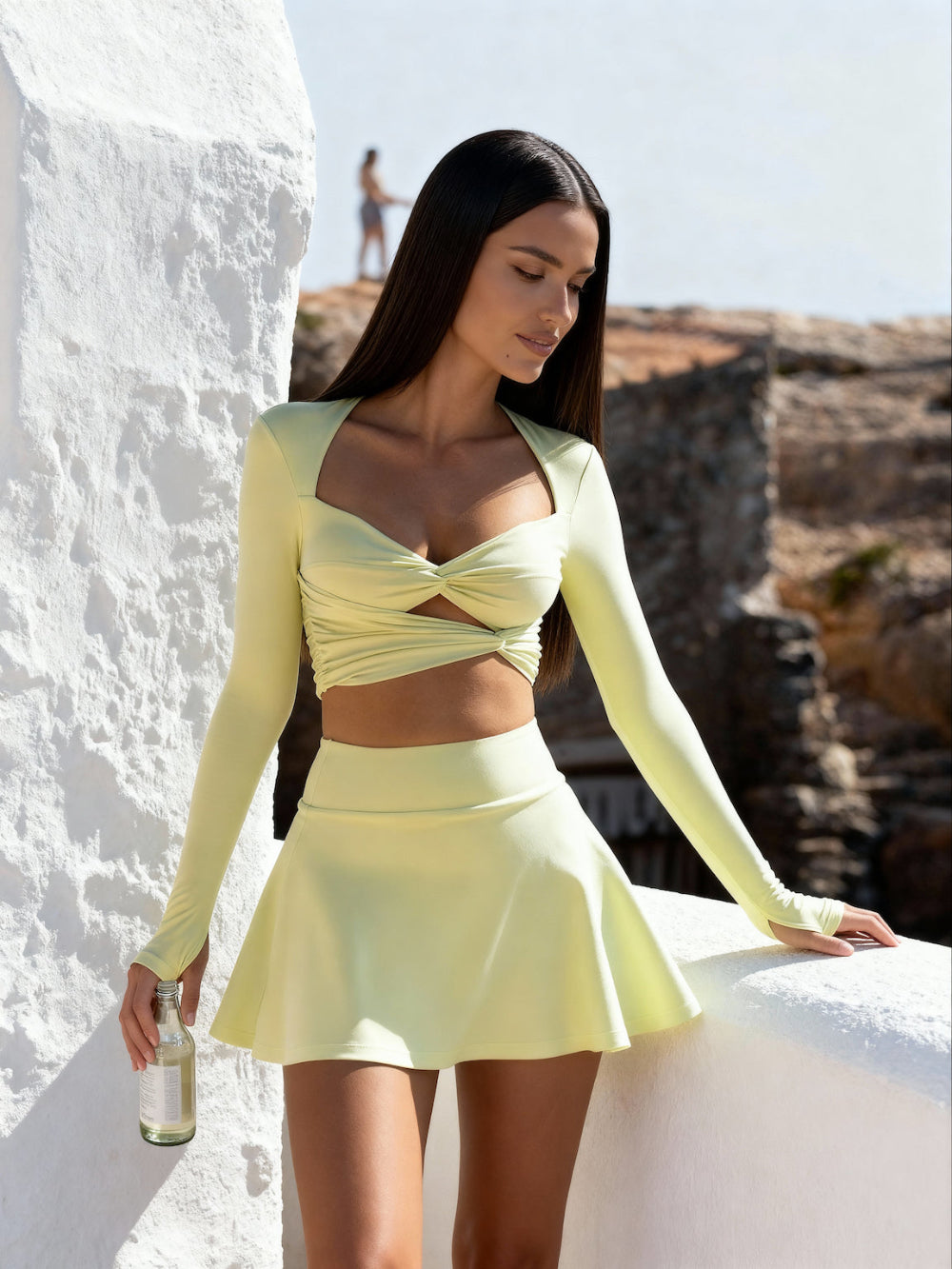 Woman in a light yellow outfit standing against a white wall with a scenic background.